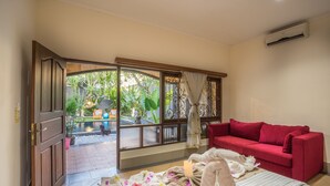 A room with a red sofa, a wooden door leading to an outdoor area with a pool, and a view of greenery. - Villa Mia And Spa (Seminyak)