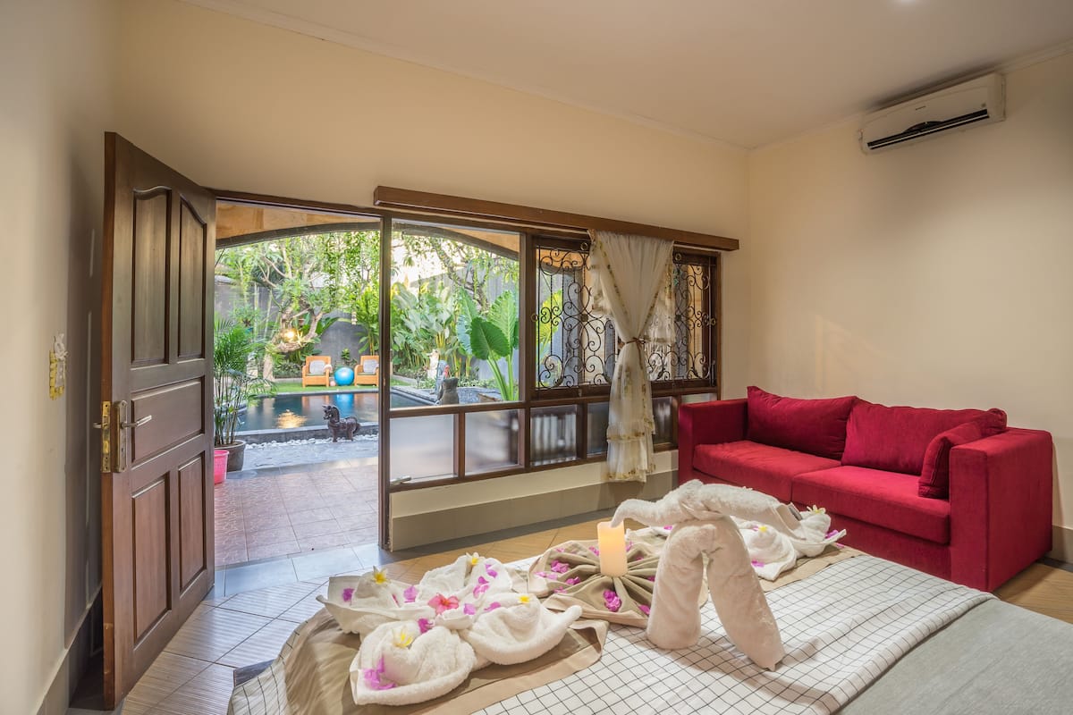 A room with a red sofa, a wooden door leading to an outdoor area with a pool, and a view of greenery.