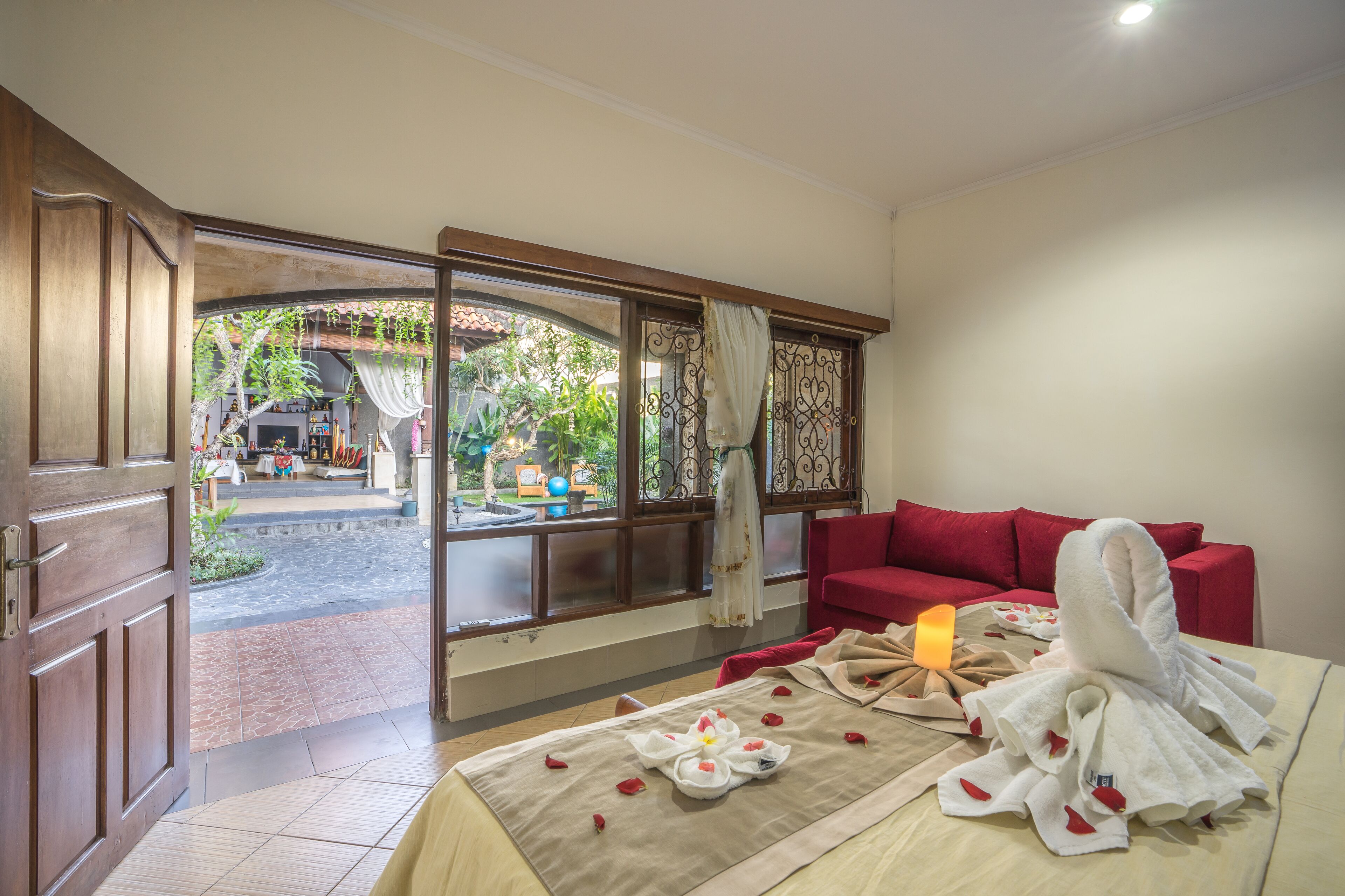 A hotel room with a bed, a red sofa, a window with a view of the garden, and a door leading outside.