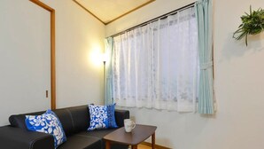A living room with a black sofa, two patterned pillows, a small wooden table, and a window with sheer curtains. - Guest House Asato (Naha)
