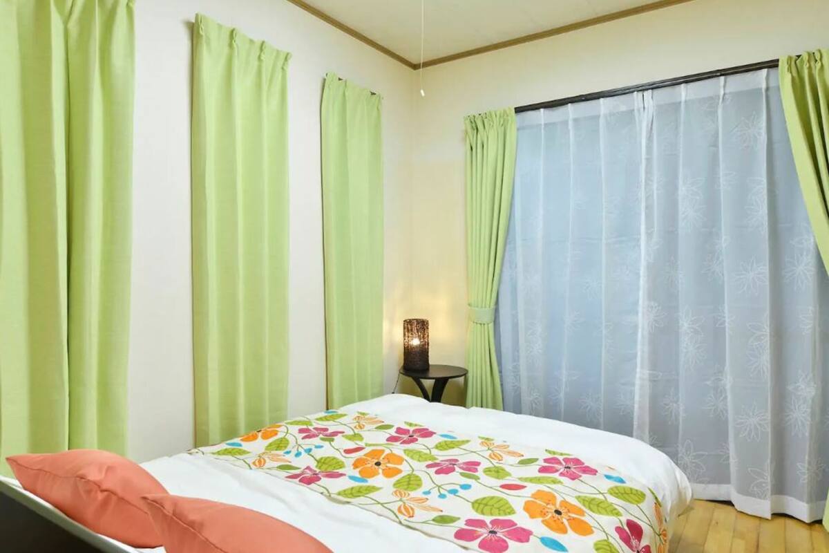 Un dormitorio con cama, cortinas verdes, una mesita de noche con lámpara y una ventana con cortinas traslúcidas.
