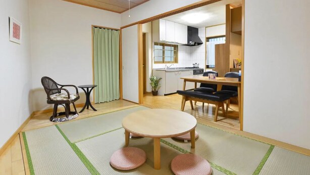 A traditional Japanese-style room with tatami flooring, a tatami mat table, a low table with a chair, and a sliding door leading to a kitchen area. - Guest House Asato (Naha)