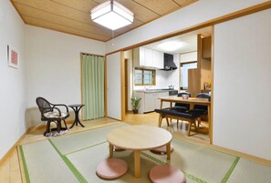 A traditional Japanese-style room with tatami flooring, a tatami mat table, a low table with a chair, and a sliding door leading to a kitchen area.