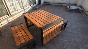 Een houten zitgedeelte buiten, met een tafel en bank, een klein opstapje naar een hoger niveau en een raam met transparante gordijnen.