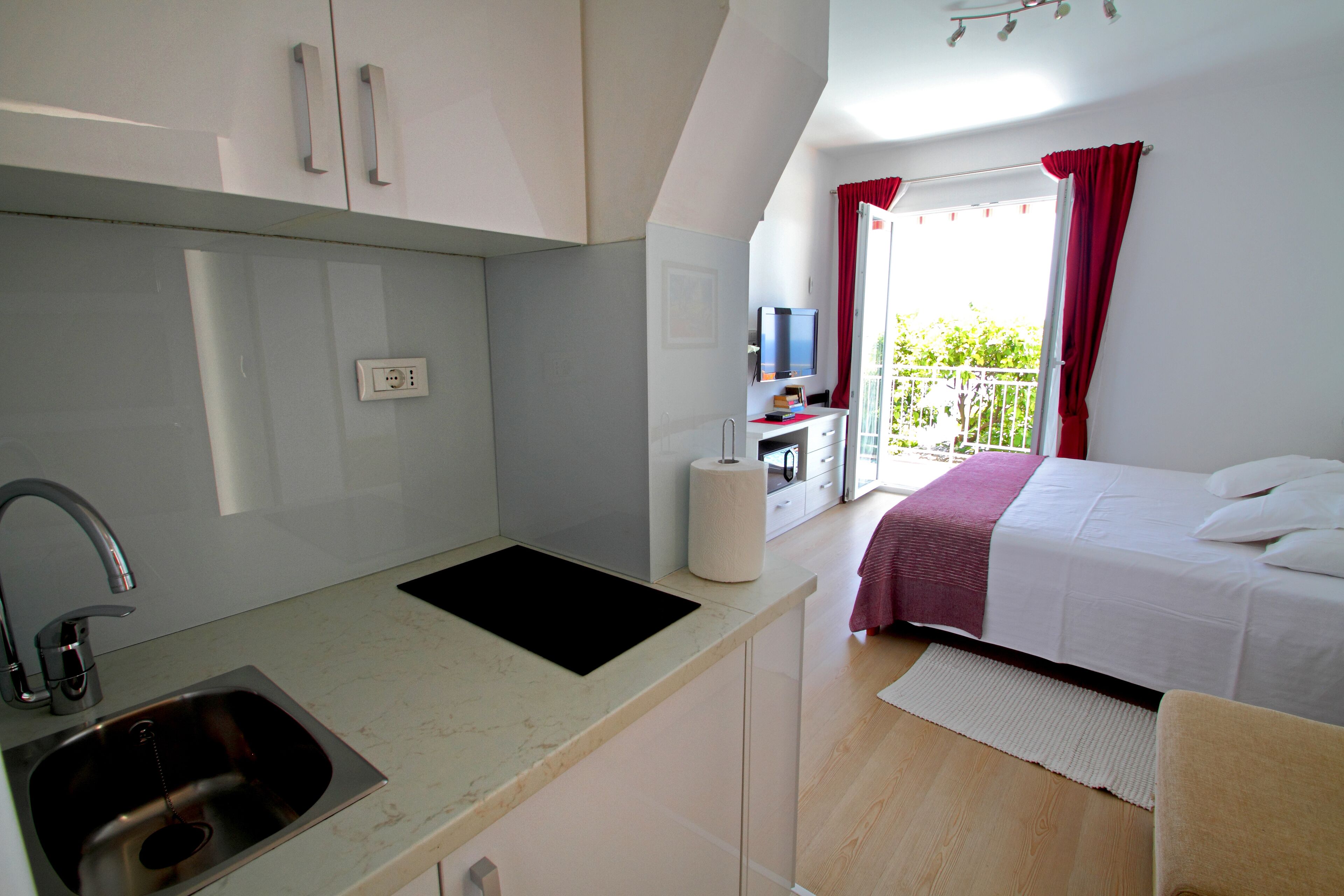A compact hotel room with a kitchenette, a bed, a TV, and a balcony with red curtains.