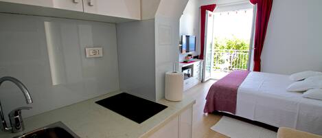 A compact hotel room with a kitchenette, a bed, a TV, and a balcony with red curtains.