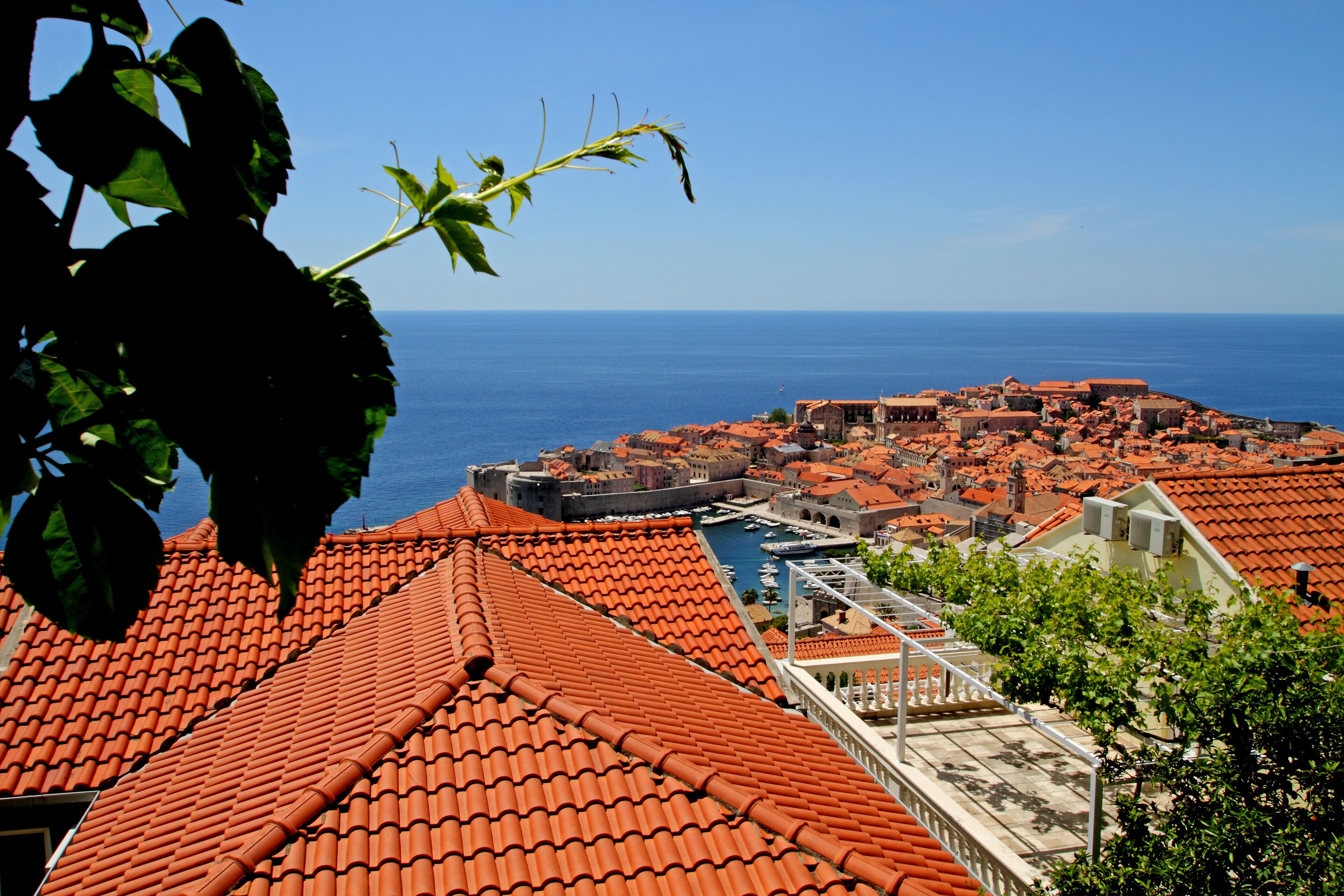 Une ville côtière aux bâtiments aux toits de tuiles rouges, dotée d’un port de plaisance et surplombée d’un ciel bleu azur.