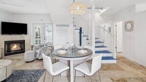 Un salon moderne avec une cheminée, un coin repas avec une table en verre, des chaises blanches et un escalier avec des marches bleues.