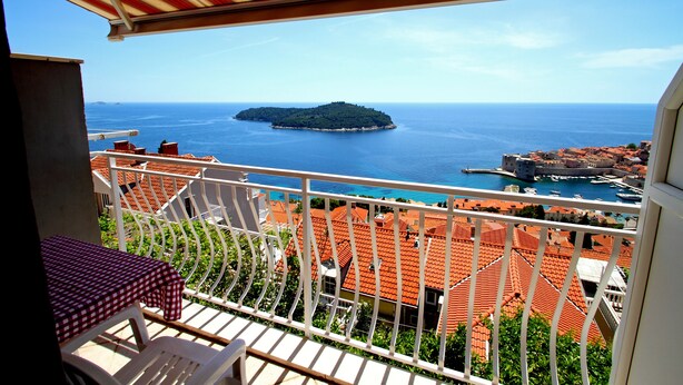 Un balcon doté d’un auvent rayé de rouge et de blanc, donnant sur une ville côtière aux toits de tuiles et sur une petite île qui se profile à l’horizon.
