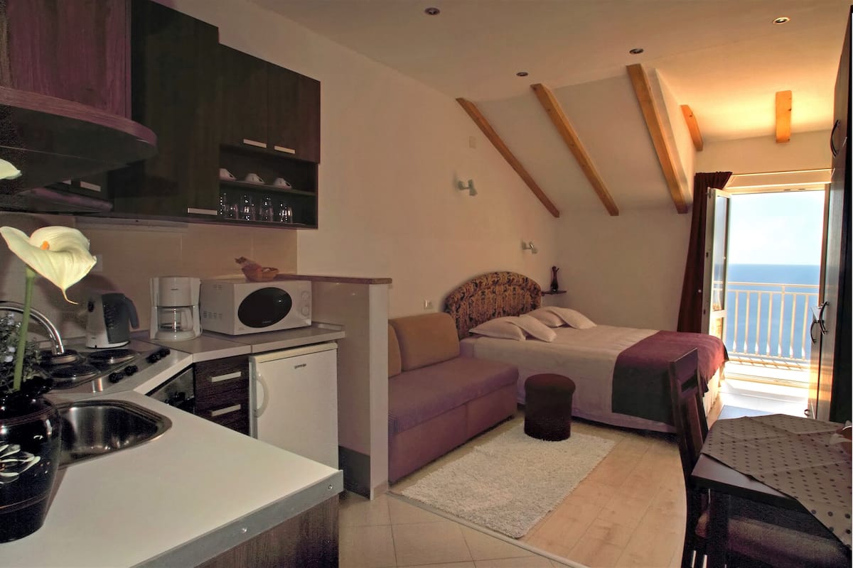 A compact room with a kitchenette, a bed with a view, a sofa, and a small table.