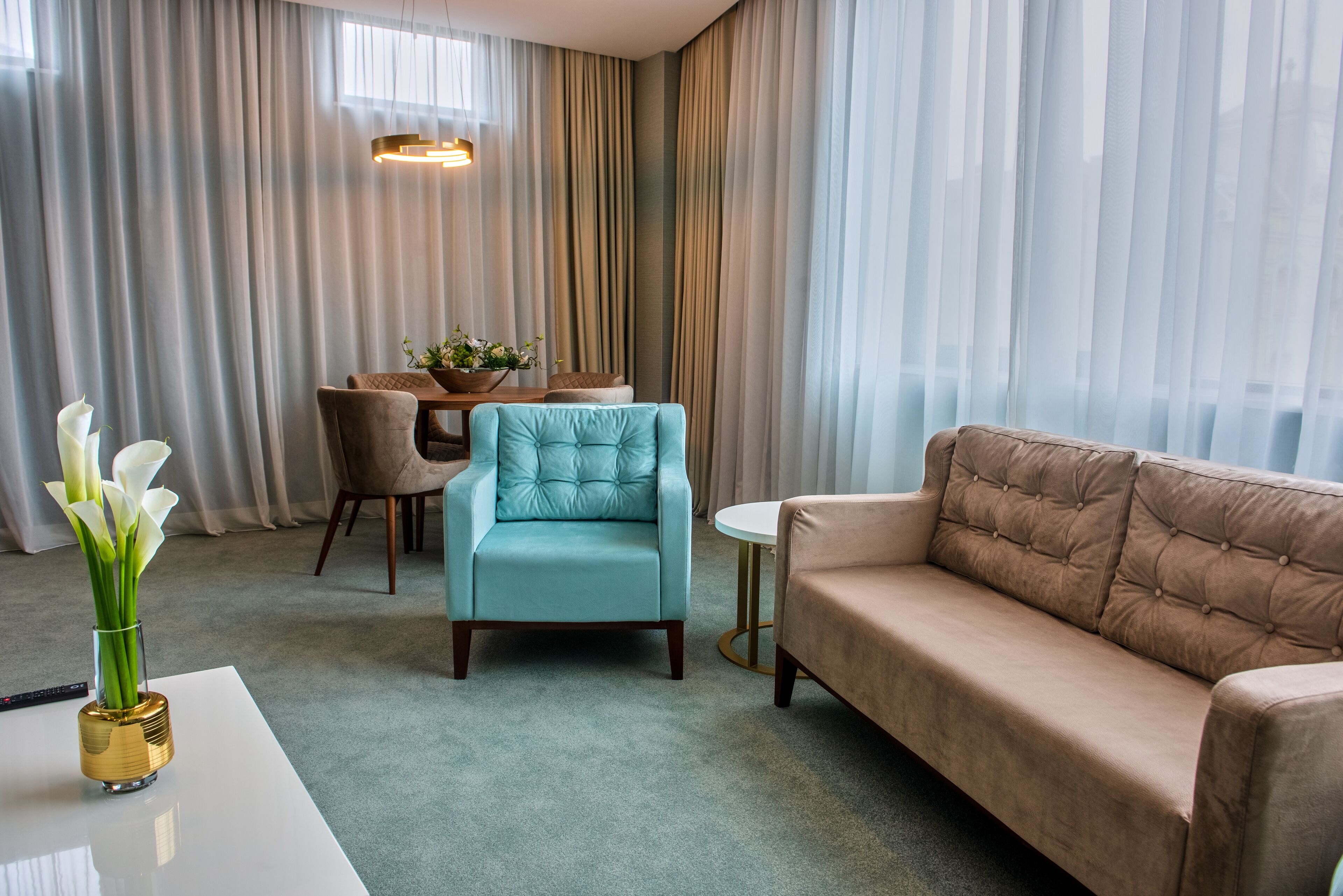 Una sala de estar moderna con un sofá, un sillón y una mesa de comedor con sillas. Hay un jarrón con flores sobre una mesa y una ventana con cortinas.