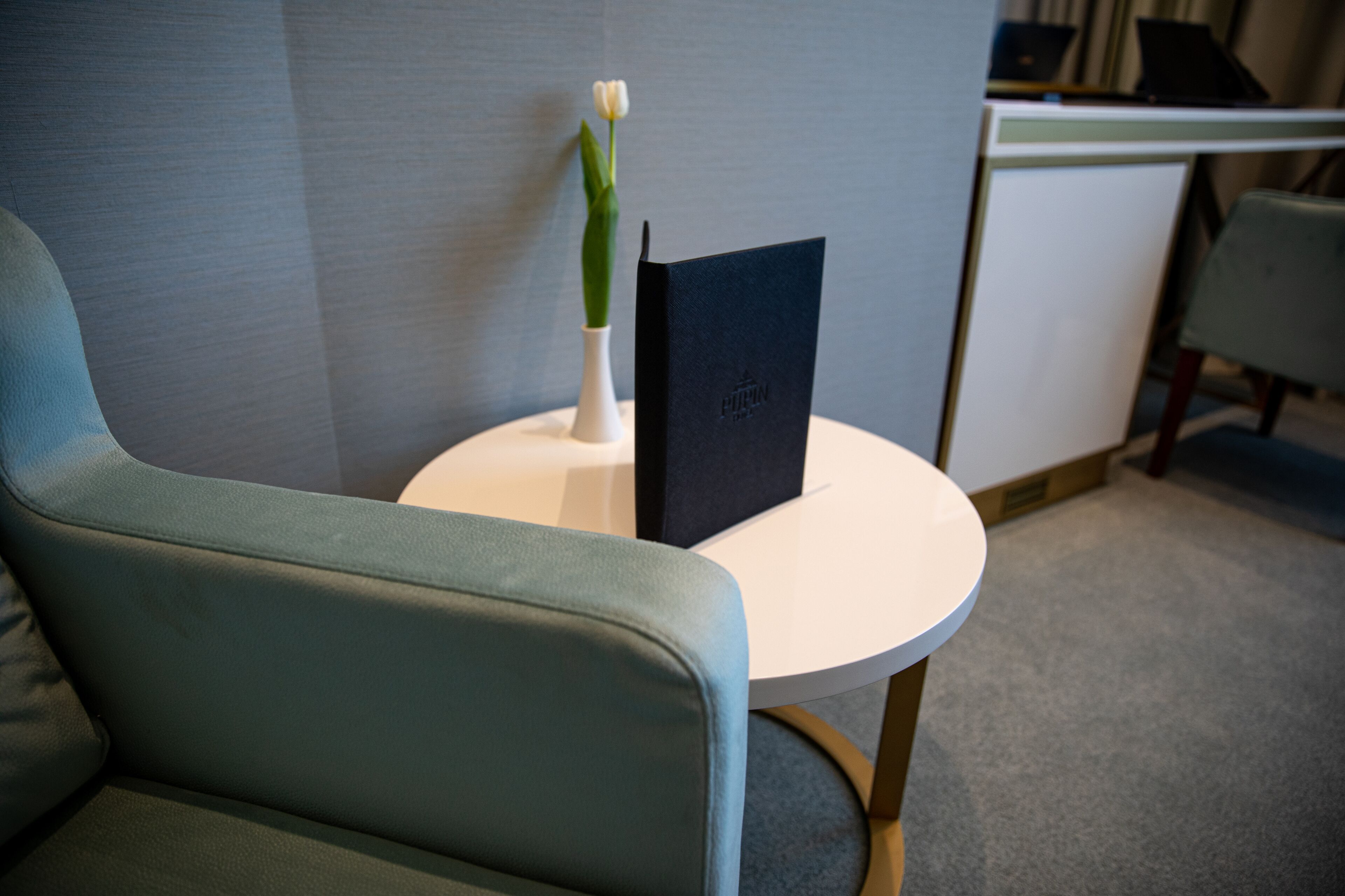 Habitación de hotel moderna con una silla tapizada de verde, una mesa redonda blanca y una libreta o carpeta negra.
