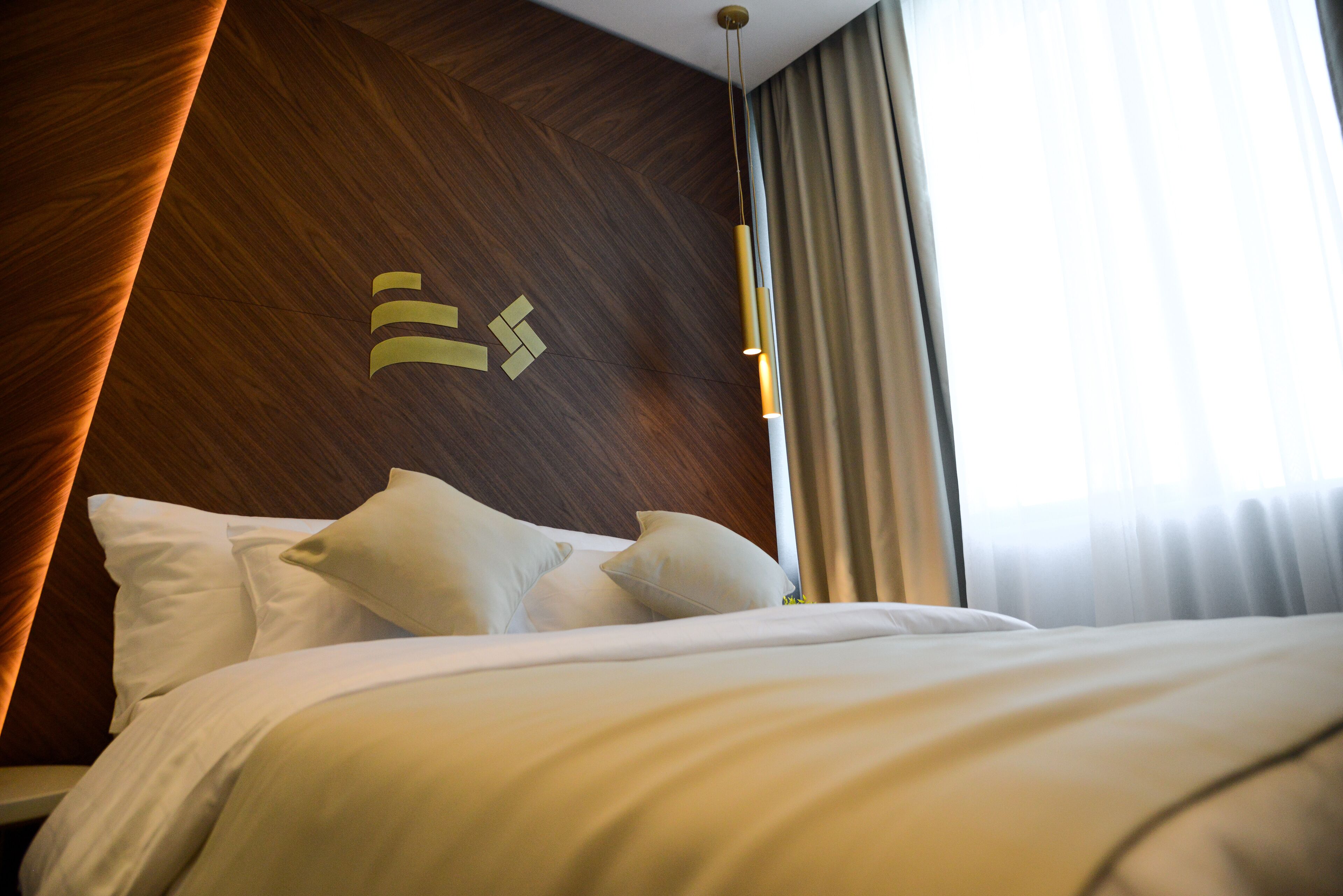 A neatly made bed with white bedding and pillows, a wooden wall with a geometric pattern, and a window with curtains.