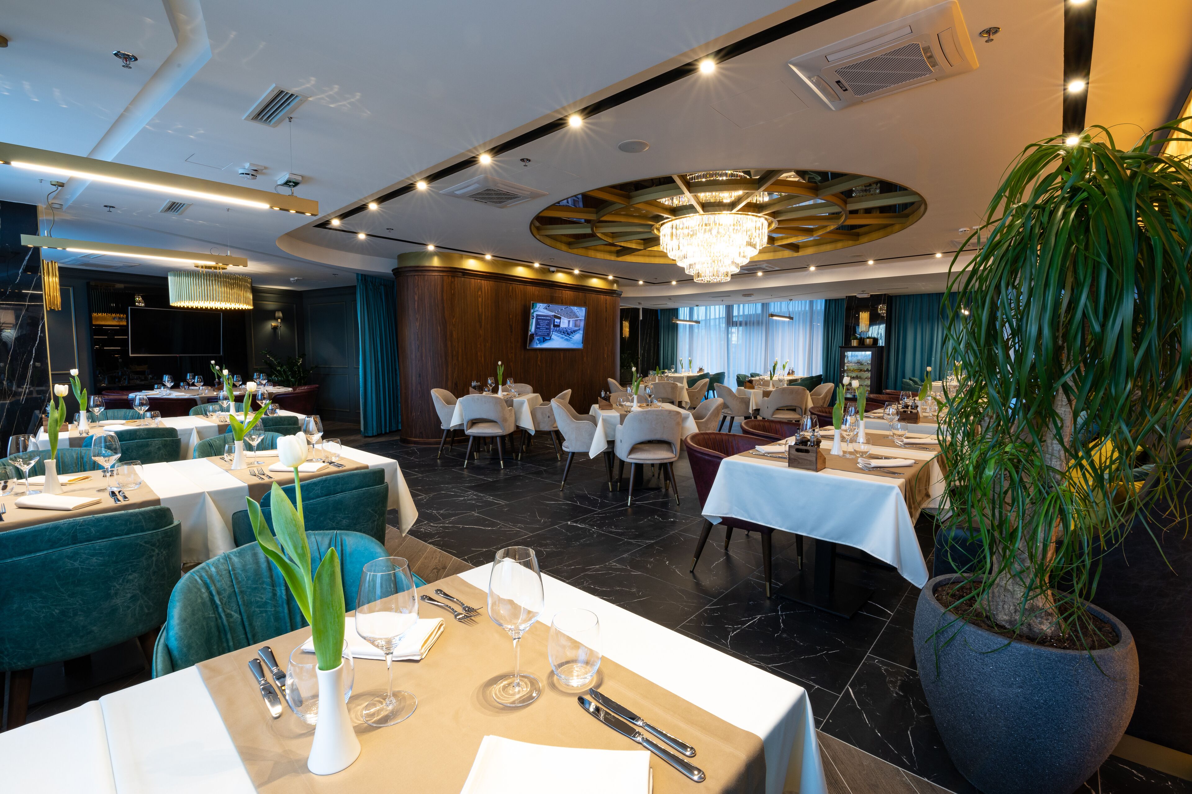 A restaurant with multiple tables set for dining, a large potted plant, and a chandelier.