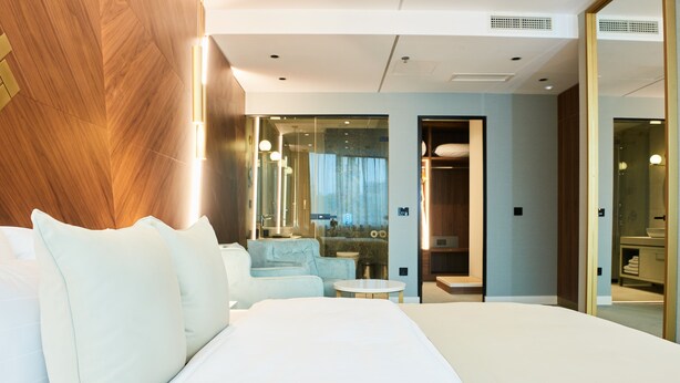 Una habitación de hotel moderna con una cama grande, una zona de estar con un sofá y una mesita, y vistas a través de una puerta de cristal.