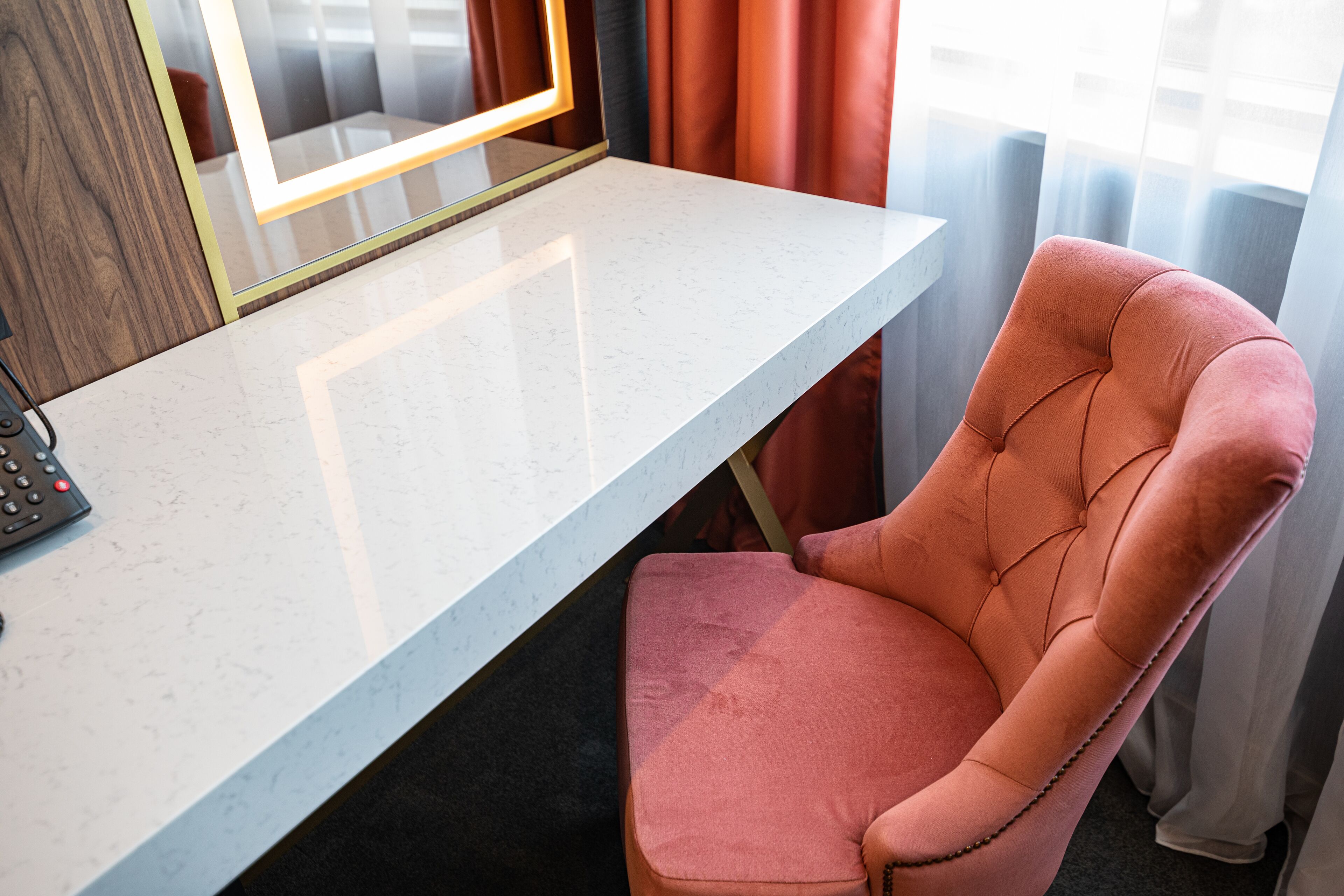 A room with a white marble desk, a pink upholstered chair, a mirror with lights, and a window with curtains.