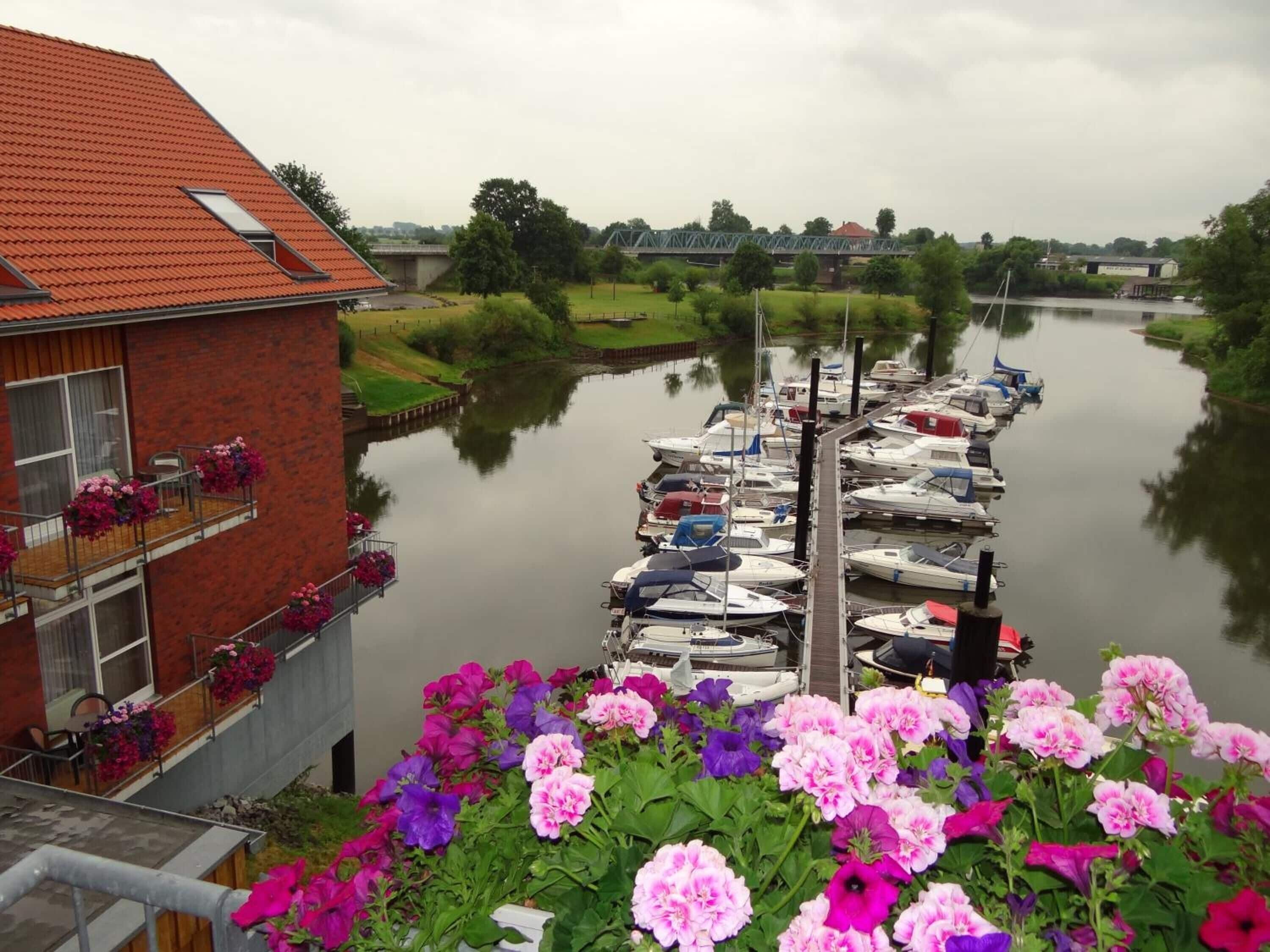 A canal with boats docked on the right, a brick building with a red roof on the left, and a row of flowers in the foreground.