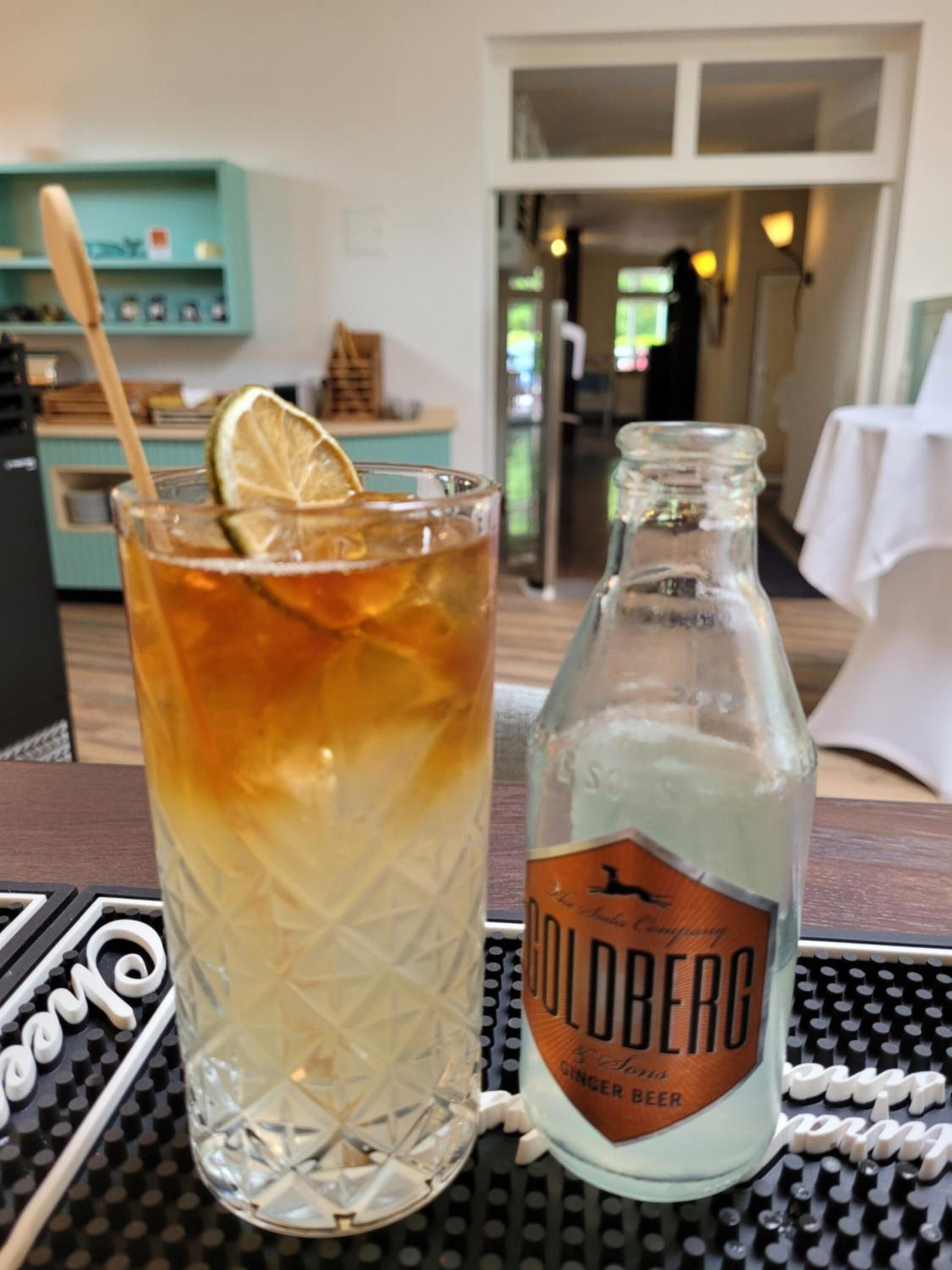 A glass of iced tea with a lemon slice, a bottle of Goldberg ginger beer, and a black and white patterned tablecloth.