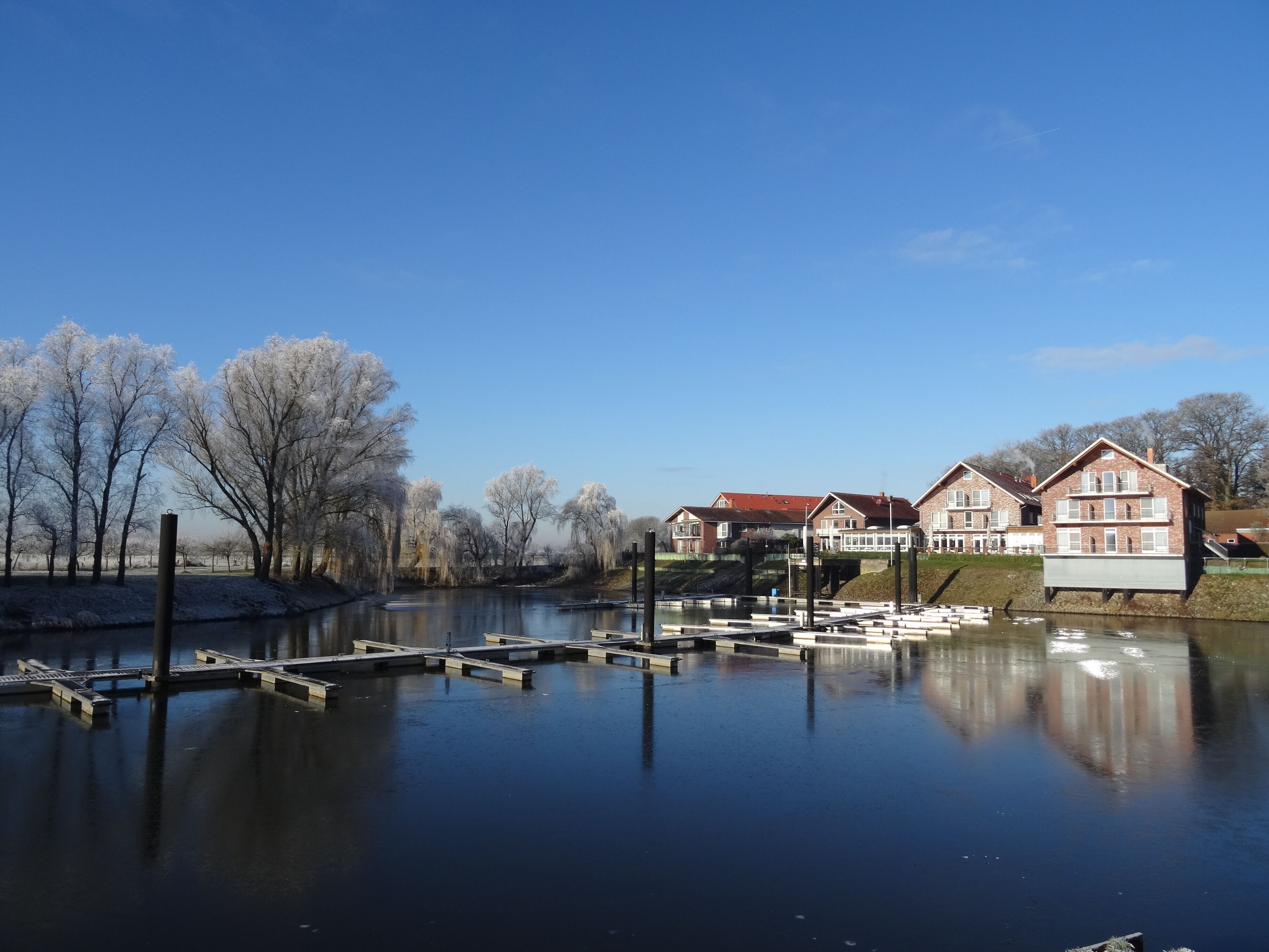 A calm river with a dock, frost-covered trees, and residential buildings in the background.