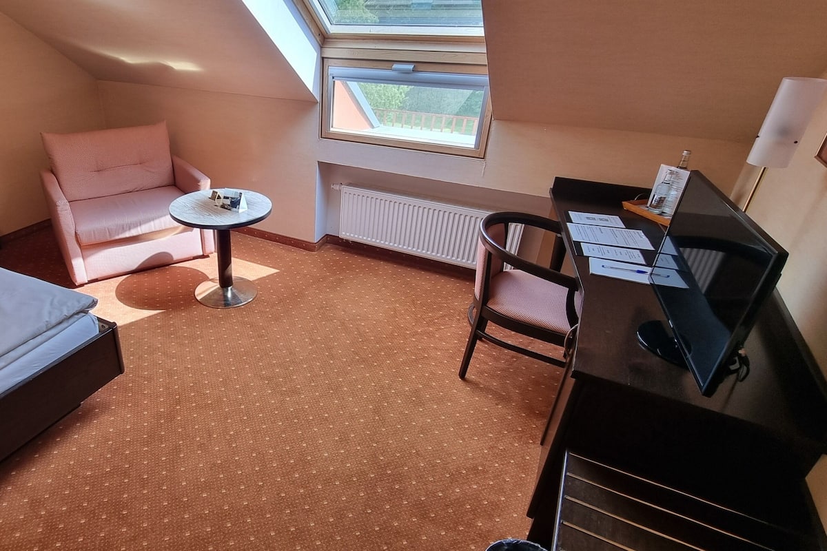 A compact hotel room with a bed, a small desk, a chair, a pink armchair, and a window providing natural light.