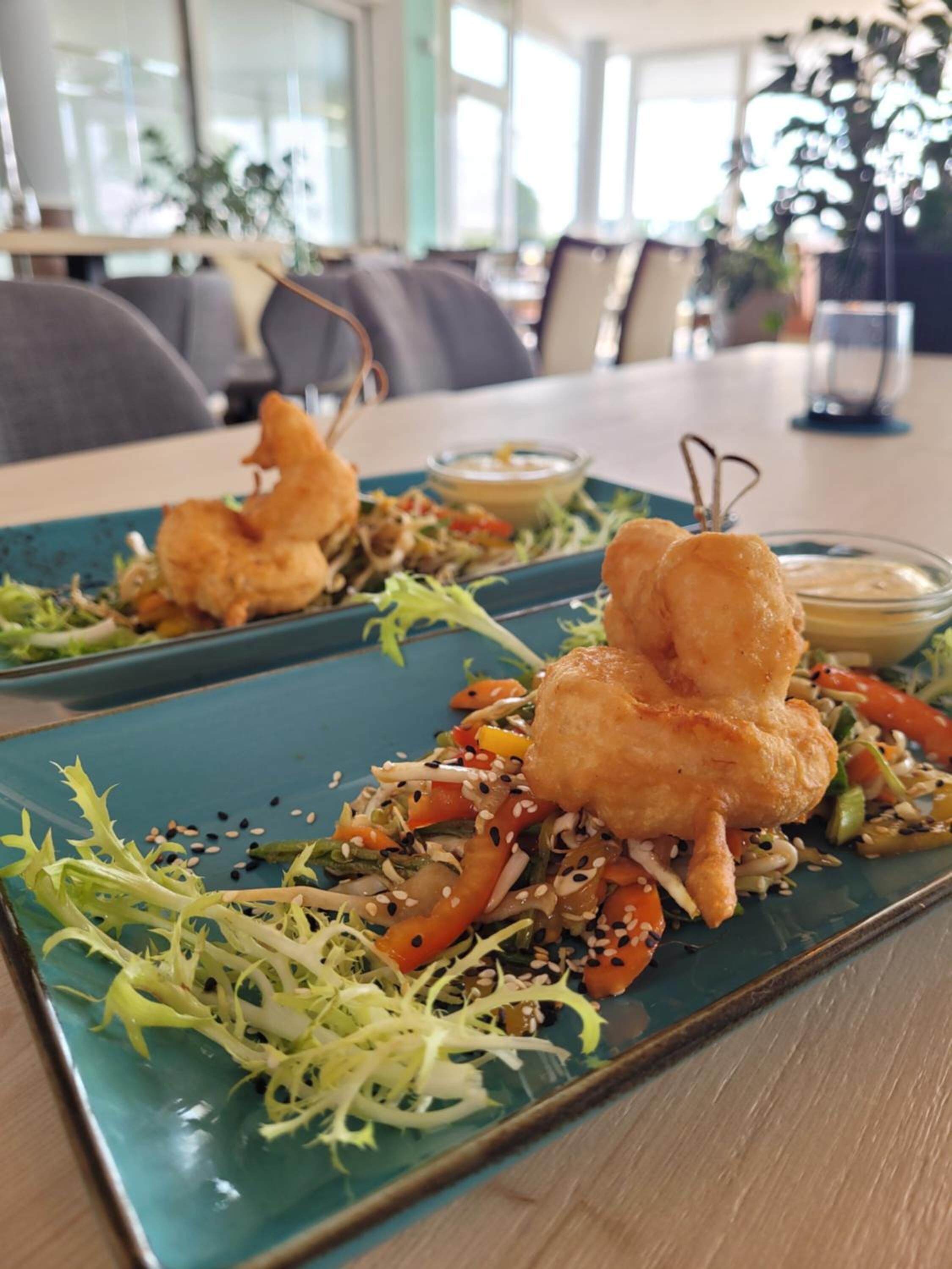 Una mesa de comedor con un plato azul que contiene una ensalada y dos porciones de comida frita, posiblemente alitas de pollo, junto con salsa para mojar.
