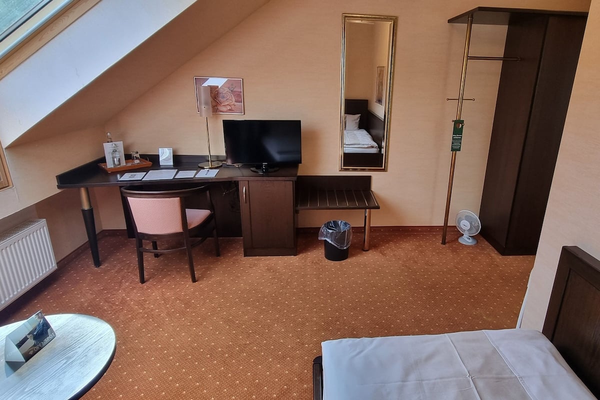 A compact hotel room with a sloped ceiling, a desk with a computer, a chair, a small table with a nameplate, a bed, a mirror, and a radiator.