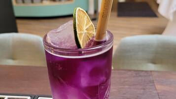 Ein violettes Getränk mit Limettenscheibe und Trinkhalm in einem strukturierten Glas, auf einem schwarzen Tisch mit weißer Schrift.