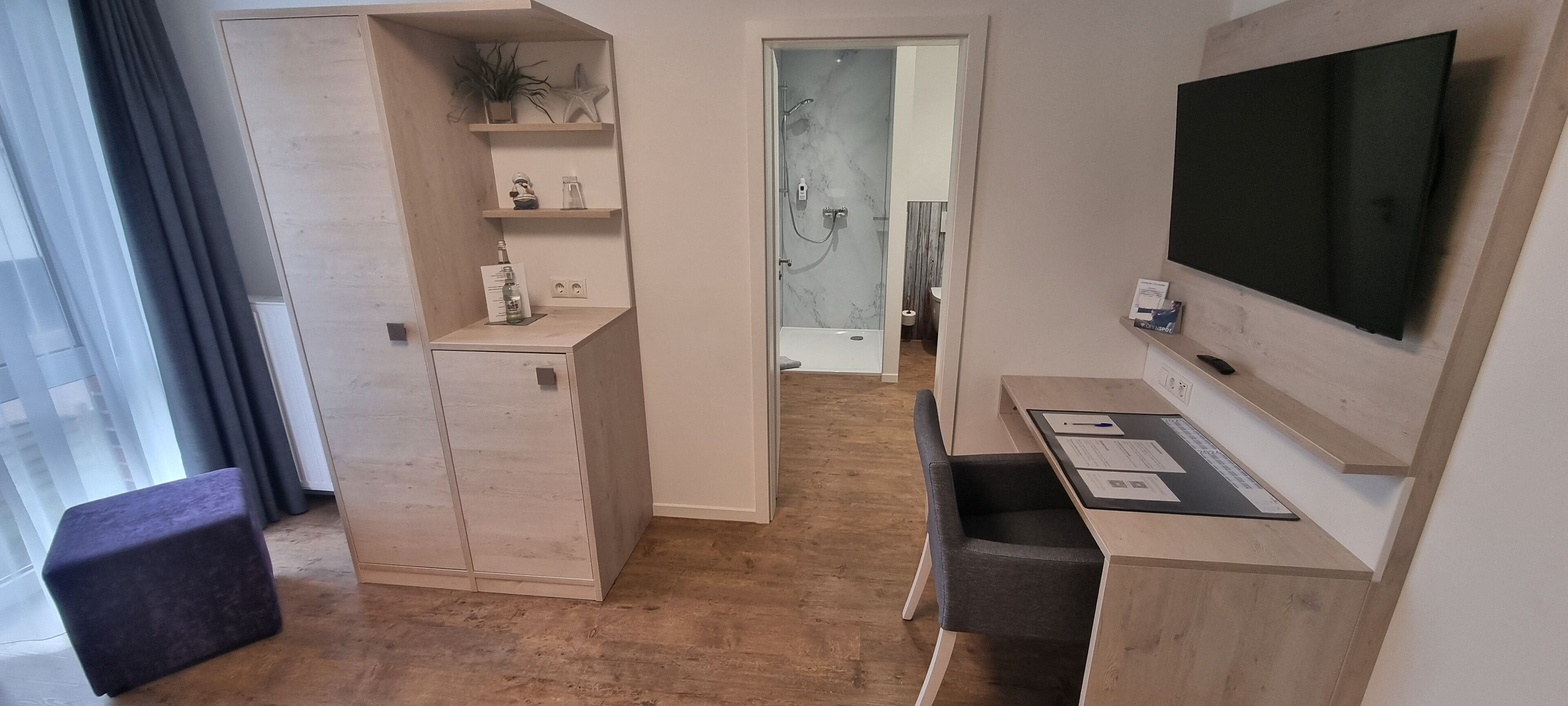 Habitación de hotel moderna con suelo de madera, televisor montado en la pared, escritorio con ordenador, mueble pequeño y estante con adornos.