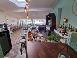 A bar area with a wooden counter, stools, and a variety of bottles and glasses. There are tables and chairs in the background, and a person is visible at the counter.