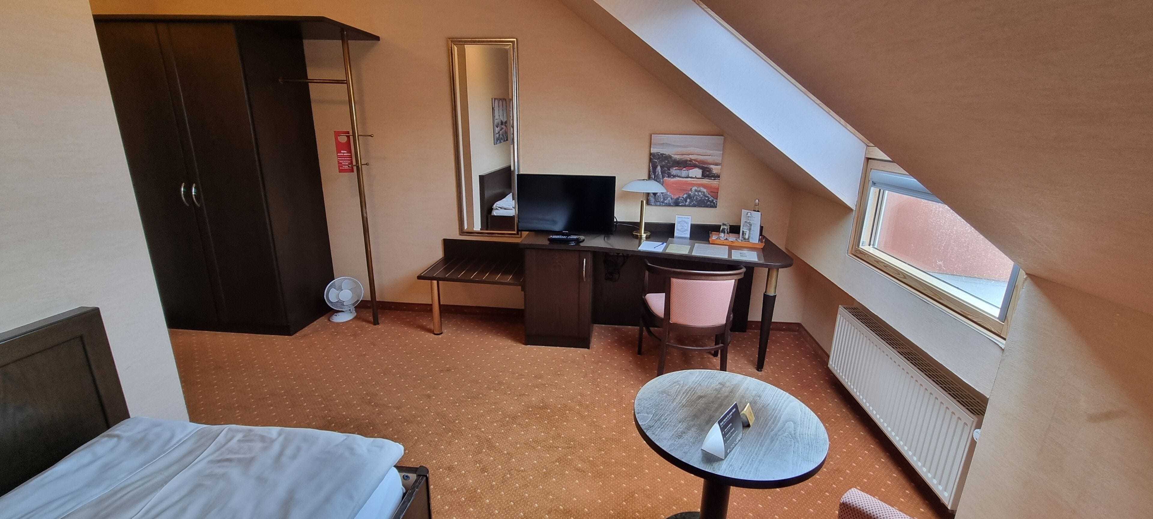 Habitación de hotel con cama, escritorio, computadora, silla, mesita y espejo, además de una ventana.