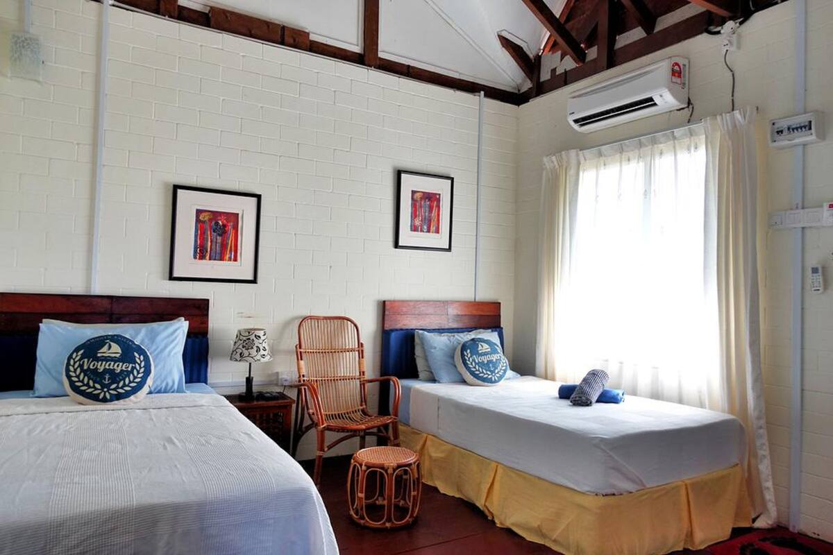 A room with two beds, a wicker chair, and a small stool. There are framed pictures on the wall, an air conditioning unit, and a window with curtains.