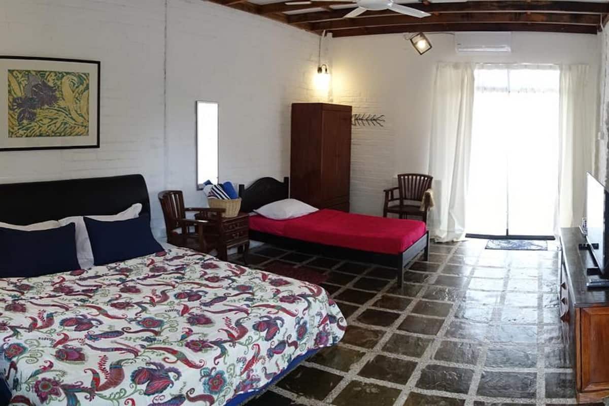 A room with a large bed, a smaller bed, a desk with a chair, a television, and a window with curtains.