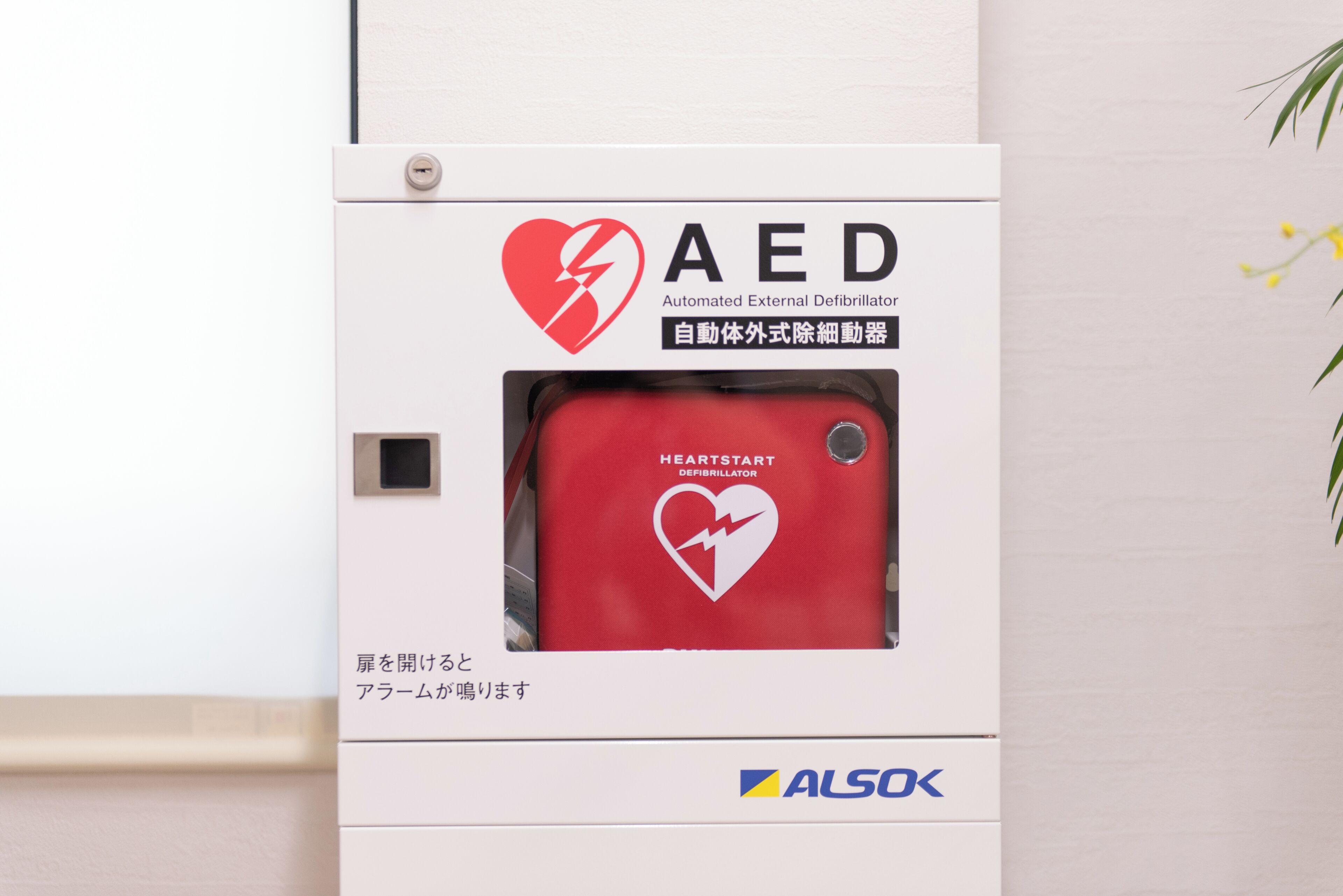 Un desfibrilador externo automático (DEA) montado en la pared, con carcasa roja y símbolo de corazón, etiquetado 'HEARTSTART DEFIBRILLATOR'.