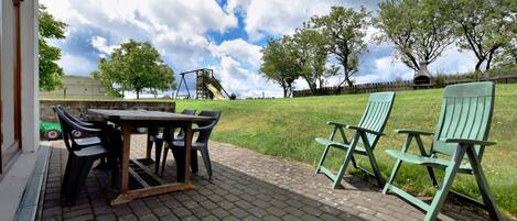 A patio with a table and chairs, a swing set, and a grassy area with trees.