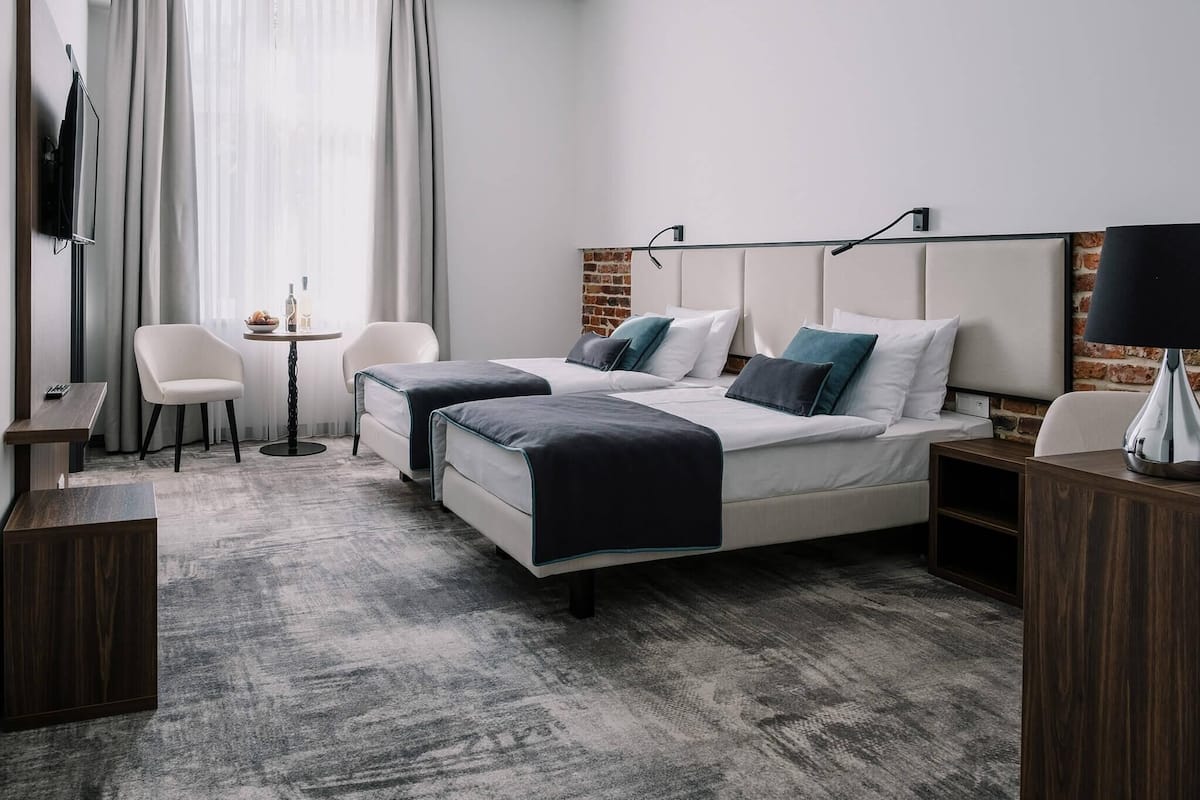 Quarto de hotel moderno com uma cama grande, duas mesas de cabeceira, uma pequena mesa redonda com uma luminária, uma TV fixada na parede e vista para a cidade através de grandes janelas.