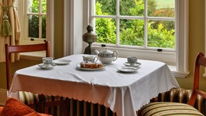 Une table à manger dressée avec un service à thé et des pâtisseries, placée près d’une fenêtre donnant sur un paysage verdoyant.