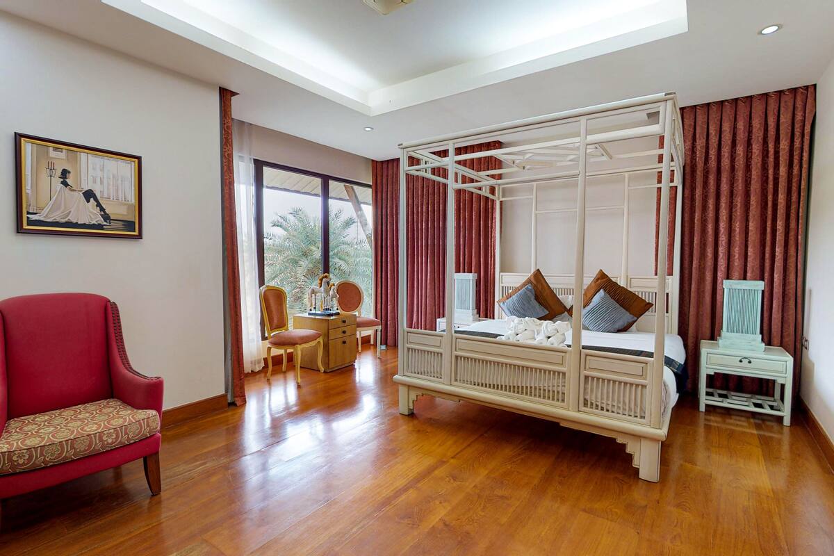 A spacious room with a four-poster bed, a red armchair, a wooden desk, and a large window with curtains.