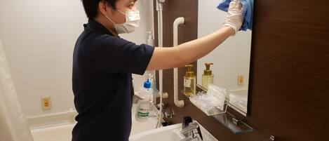 A person cleaning a mirror in a bathroom with a sink, toiletries, and a towel rack.