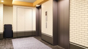 Ein Aufzug mit digitaler Anzeige, ein Gepäckwagen und ein gemustertes Teppich im modernen Eingangsbereich mit Backsteinwänden und Holzeinzelungen an der Decke.