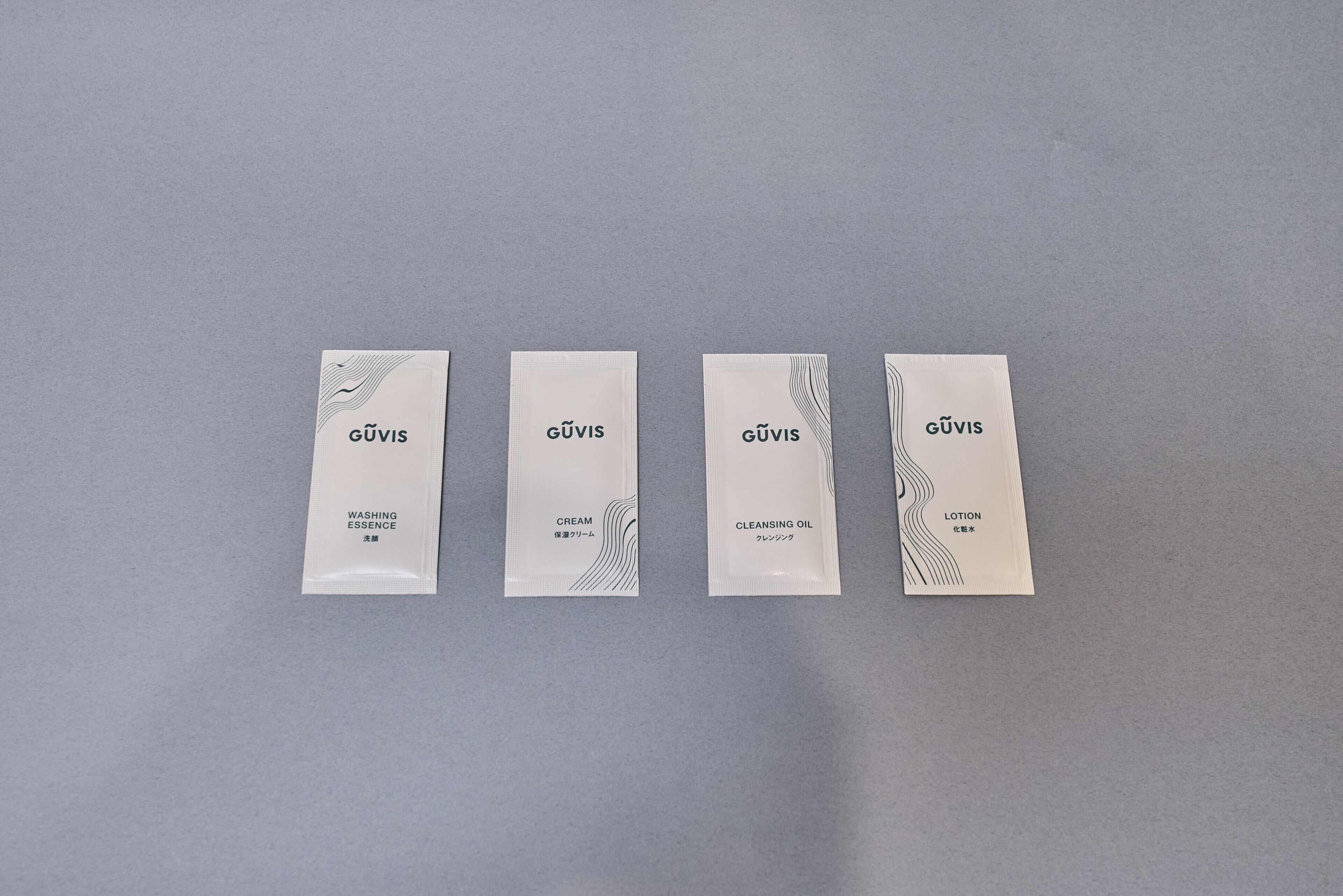 Cuatro paquetes blancos rectangulares con el nombre de marca 'GÜVIS' y los nombres de producto 'WASHING ESSENCE', 'CREAM', 'CLEANSING OIL' y 'LOTION' sobre un fondo gris.