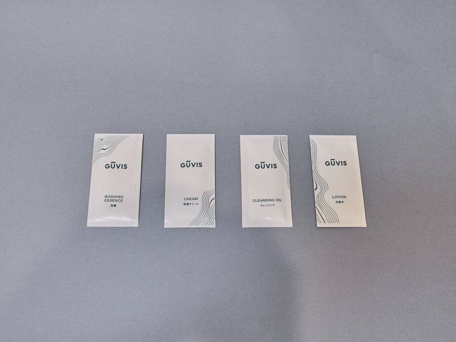 Cuatro paquetes blancos rectangulares con el nombre de marca 'GÜVIS' y los nombres de producto 'WASHING ESSENCE', 'CREAM', 'CLEANSING OIL' y 'LOTION' sobre un fondo gris.