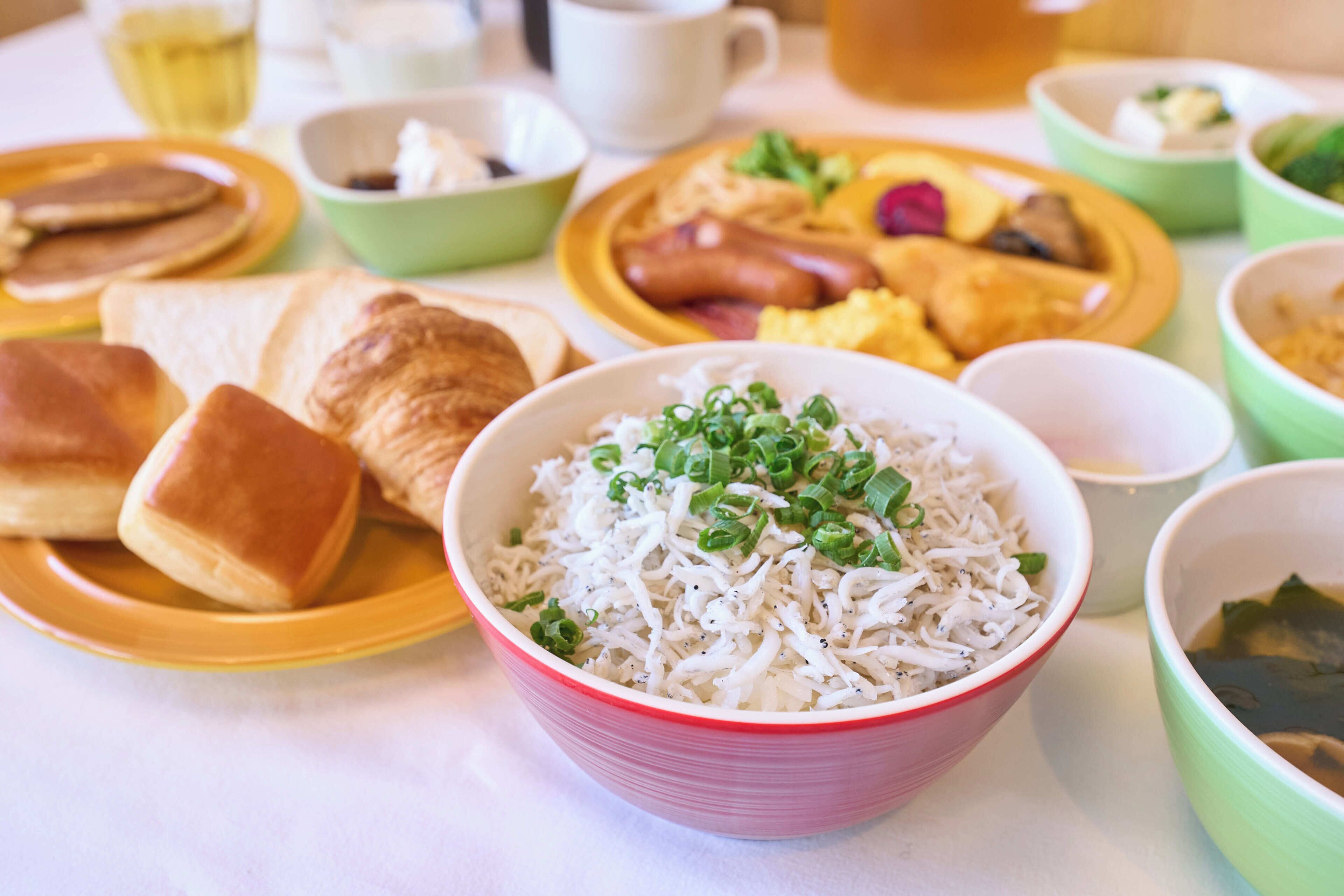 A table with various dishes including a bowl of rice with green onions, pancakes, sausages, and scrambled eggs.