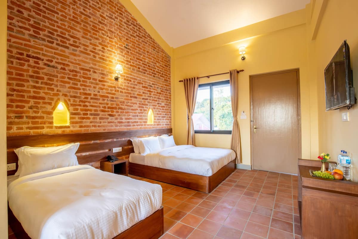 Habitación de hotel con dos camas, una pared de ladrillo, una ventana con cortinas, una puerta de madera y un pequeño escritorio con un frutero.