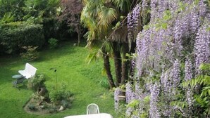 Un exuberante jardín con una mesa y sillas blancas, rodeado de diversas plantas y una enredadera floreciente morada.