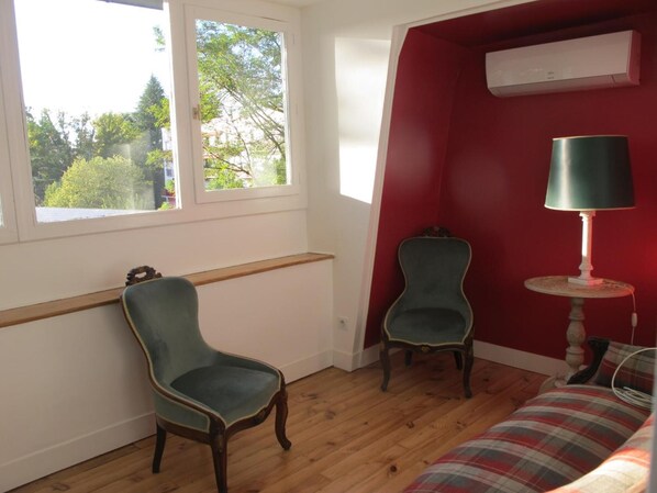 A room with a wooden floor, a red accent wall, a window with a view of trees, a green chair, a small table with a lamp, and a plaid sofa.