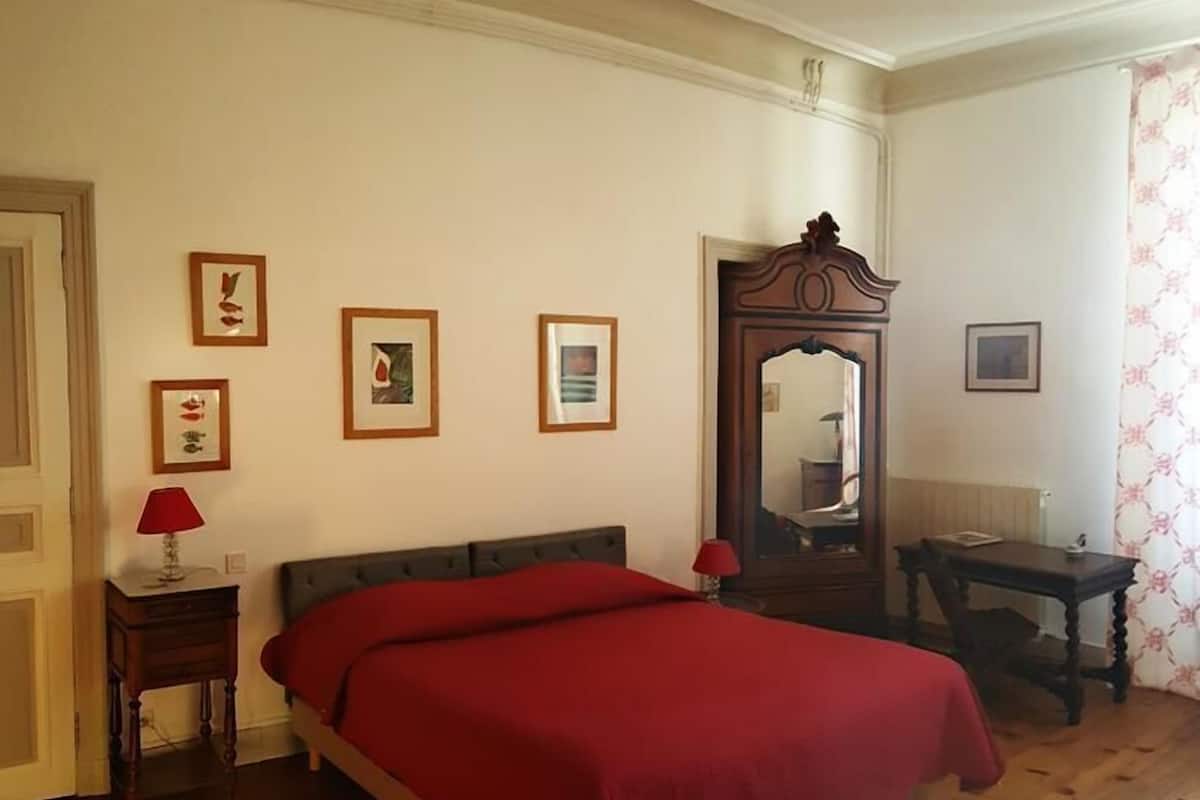 Un dormitorio con piso de madera, una cama con cubrecama rojo, una mesita de noche con lámpara, un espejo y cuadros enmarcados en la pared.