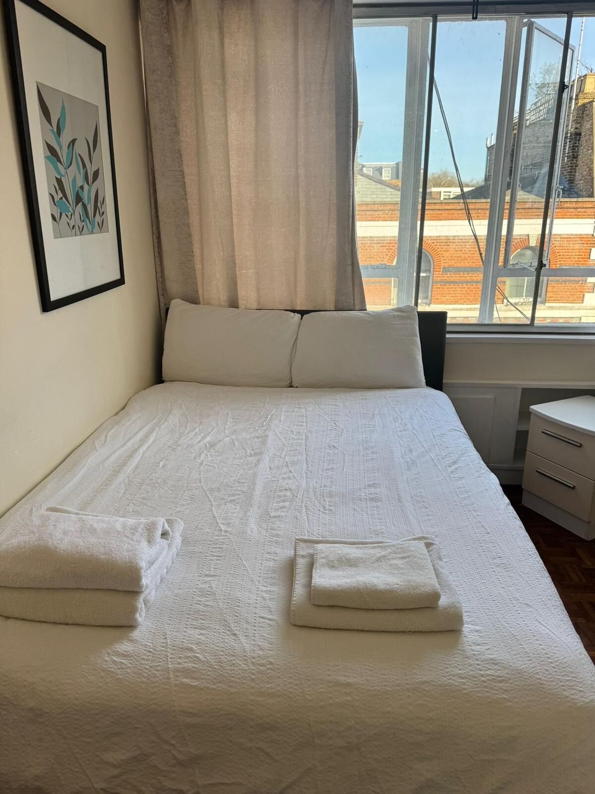 Una cama bien tendida con dos toallas dobladas, un cuadro en la pared y una ventana con vistas a edificios.