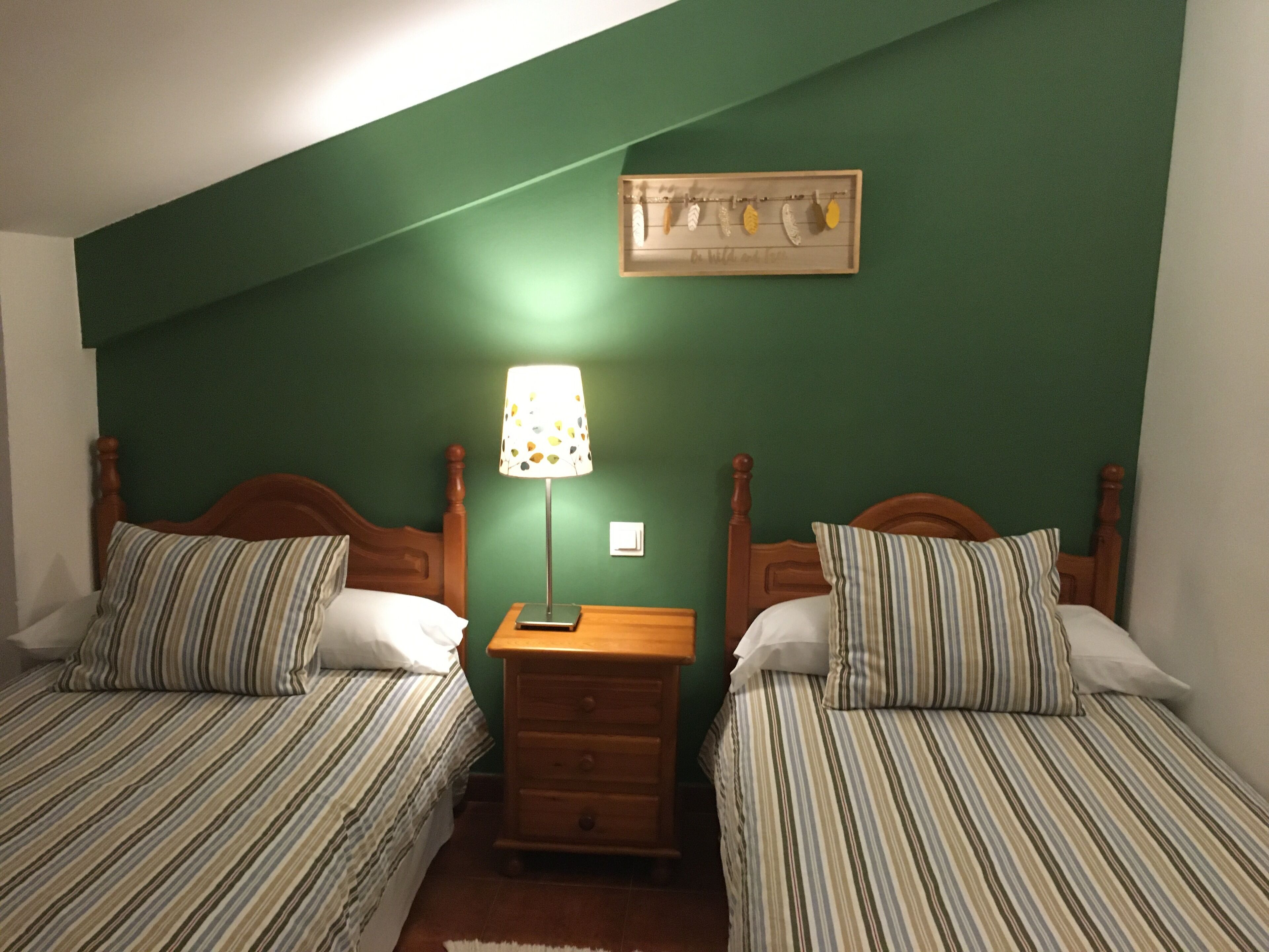 A room with two single beds, a nightstand with a lamp, and a wall-mounted shelf with decorative items.