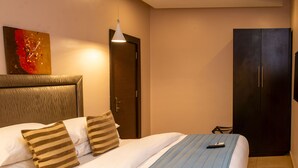 A hotel room with a bed, a wooden headboard, a painting on the wall, a wardrobe, and a remote control on the bed. - Ace Premier Hotel (Abuja)