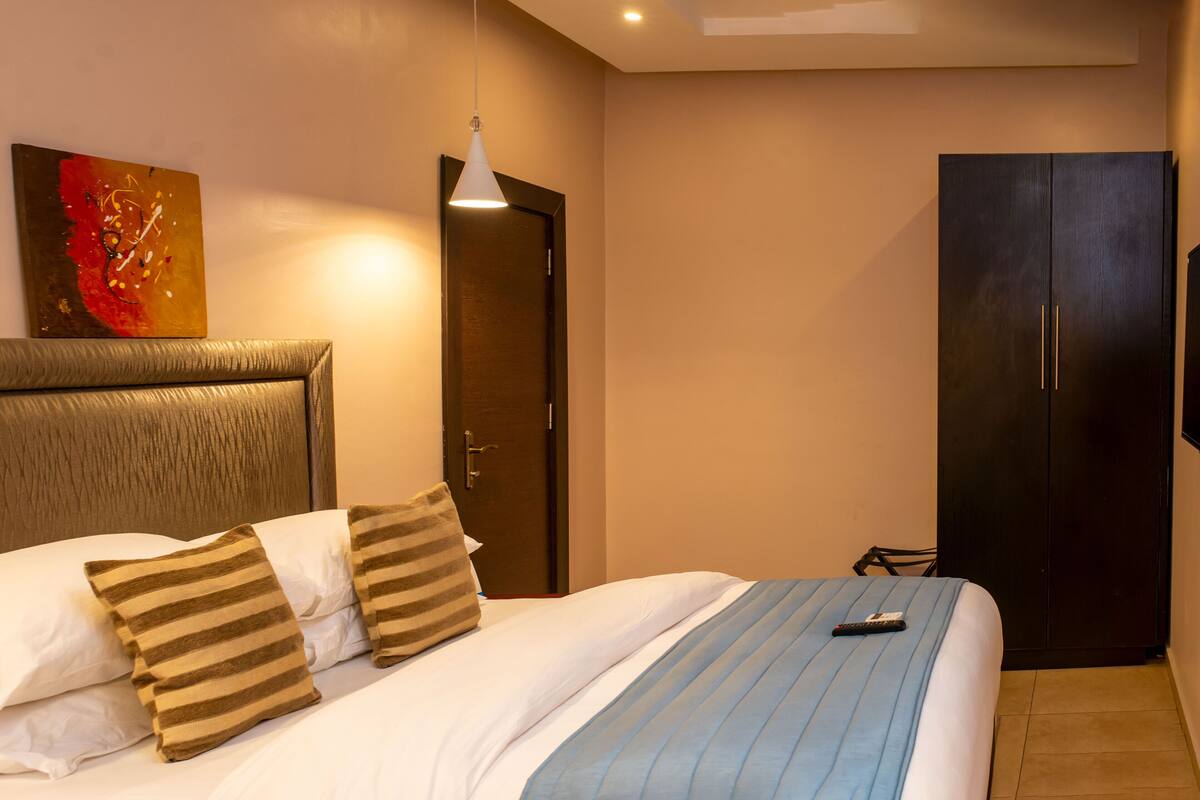 A hotel room with a bed, a wooden headboard, a painting on the wall, a wardrobe, and a remote control on the bed.